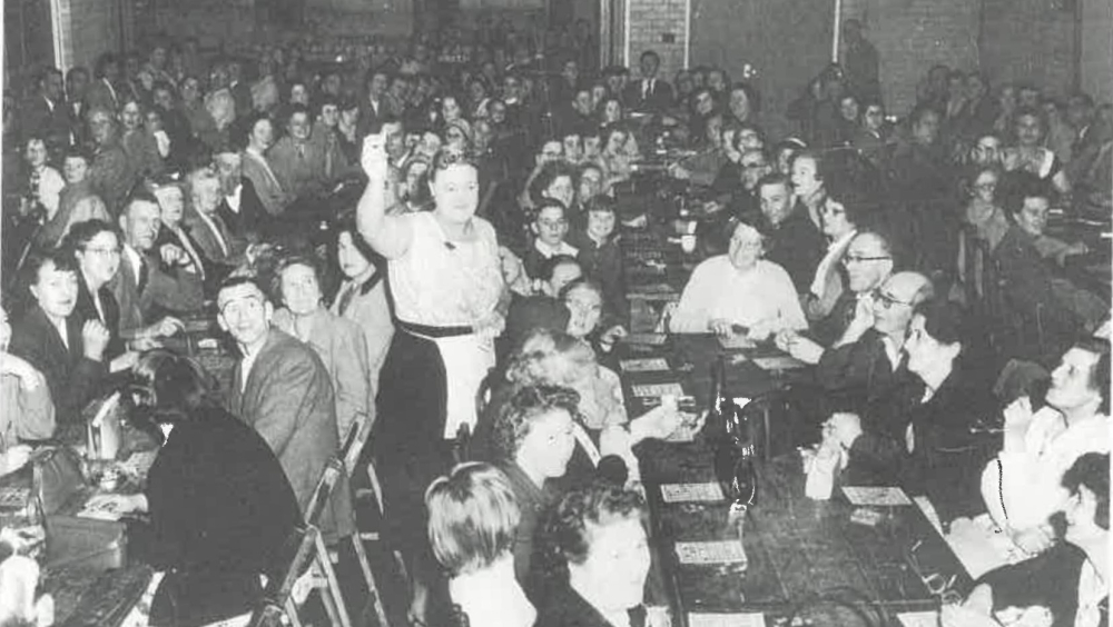 A very full room of people staying Bingo in Rose Hill. In the middle is a woman holding up a ticket.