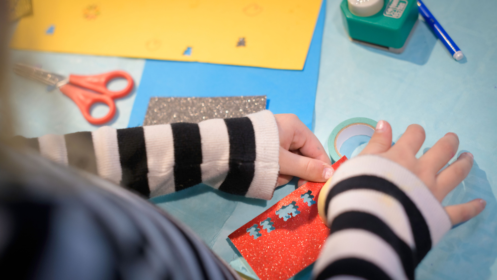  A child working on a candle holder, decorating the cardboard with glittery cut-outs in the shape of an Christmas tree. 