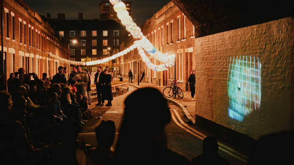An artist film is projected onto the side of a railway arch, a group of people are watching and an illuminated trail of paper lanterns runs down the street.