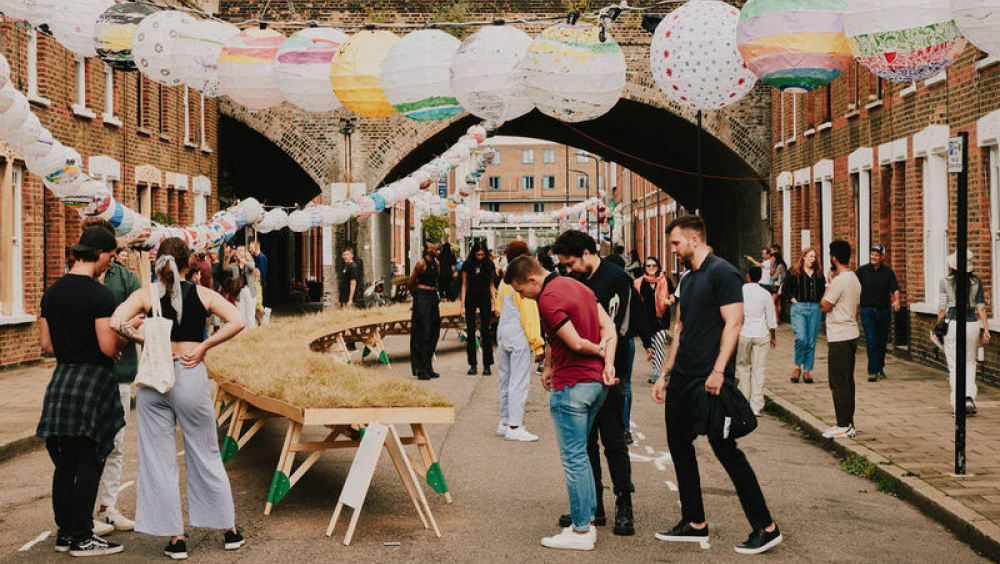 A view looking down Beck Road, towards the railway arch, the Victorian terrace houses running parallel to each other either side of the frame. Between them, Beyond Beck Road visitors are looking at the architecture, at hanging paper lanterns overhead, and at Luke Deering's art installation 'Reclaim the Commons': a meadow elevated on a tabletop running off into the distance. 