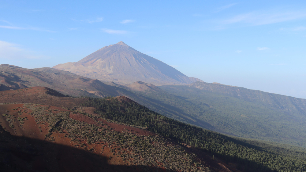 This panoramic view of Mount Teide showcases its majestic form rising above the diverse terrain of Tenerife. In the distance, a hazy veil hangs over the landscape, a telltale sign of the calima, a Saharan dust phenomenon that often affects the Canary Islands. Despite the atmospheric effect, the volcano's grandeur remains undeniable, a testament to its enduring presence on the island.