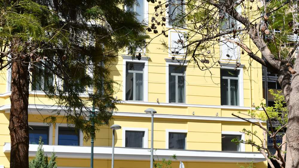 Morning or early evening hour, shiny day. In the foreground there is green grass and trees in the right and left of the photo frame. In the background there is a 3-floor neoclassical building of a light-yellow color. Part of the left side of the building is hidden by one of the trees a tree in the foreground. On the lower part of the building, a few big 3D black letters hanged on the yellow wall shape the label “Maria Callas Museum”. Some people are passing by the street, in front of the building. Two motor