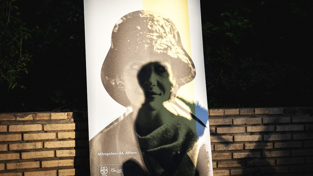  In the center of the image there is a vertical banner with a bust of Maria Callas wearing a hat, printed on white background. There is also an abstract vertical yellow thick line on the right side of the banner. On the cloth of the banner appears a shadow of a sitting man playing a wind instrument. Behind the banner there is a low horizontal brick wall, which is revealed on both sides of the banner. On the low left corner of the banner there is the Maria Callas Museum logo, as well as the address and the c
