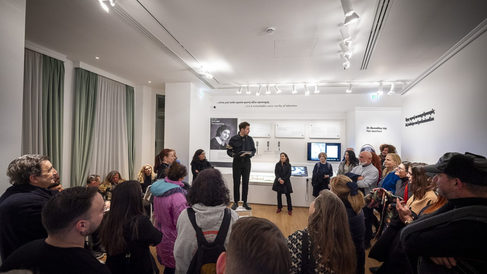An interior of an exhibition space. In the center There is a man standing on a stool, surrounded by a lot of people looking at him. On the main background there are several photos, labels and 3d letters. On the left side of the image there are two big, high windows which are covered with white curtains and in front of each curtain there is a drawn (on both sides) light-green curtain. On the right of the image there is a fake exhibition wall with a 3D lettered sentence on it.