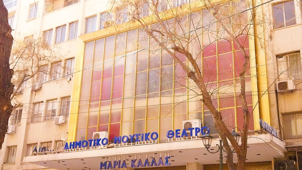 The main theme of the image is a big building on a city street. There is a big label on the upper center of the building with blue 3d letters shaping the name “CITY MUSIC THEATRE” and below that, the name “MARIA CALLAS”, both written in the Greek language. The image is taken from the opposite right side. Two cars are passing by.