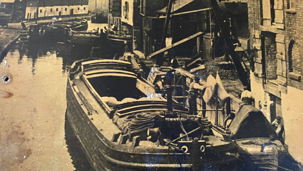 An old photo of Preston Brook wharf. In the foreground 3 men are working a crane unloading sacks from a barge into a narrowboat tied alongside. Behind them two barges are being maneuvered into a warehouse. 