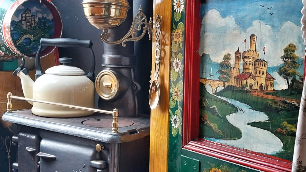 The interior of a traditional English canal boat cabin. A coal range is to the left of the image, alongside a built-in cupboard. The cabin is painted with woodgrain, trimmed in red, and has a green cupboard flap adorned with painted roses and a castle scene