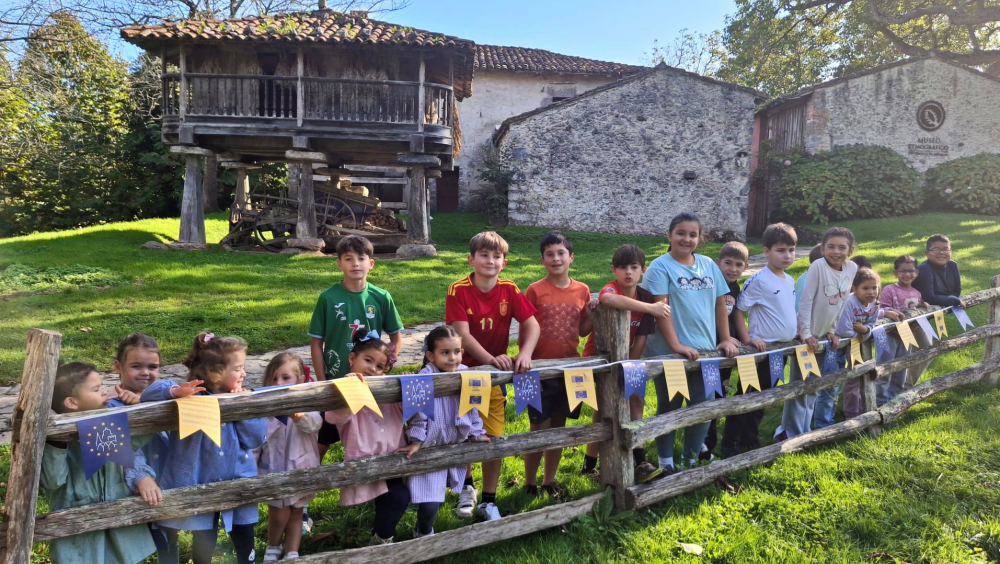 Una fila de niños y niñas está de pie junto a una valla de madera en un entorno rural verde y soleado. Muestran una guirnalda de banderillas de las JEP. Al fondo se ven construcciones tradicionales de piedra y madera, un hórreo y árboles. La escena transmite alegría y participación en una actividad comunitaria al aire libre.