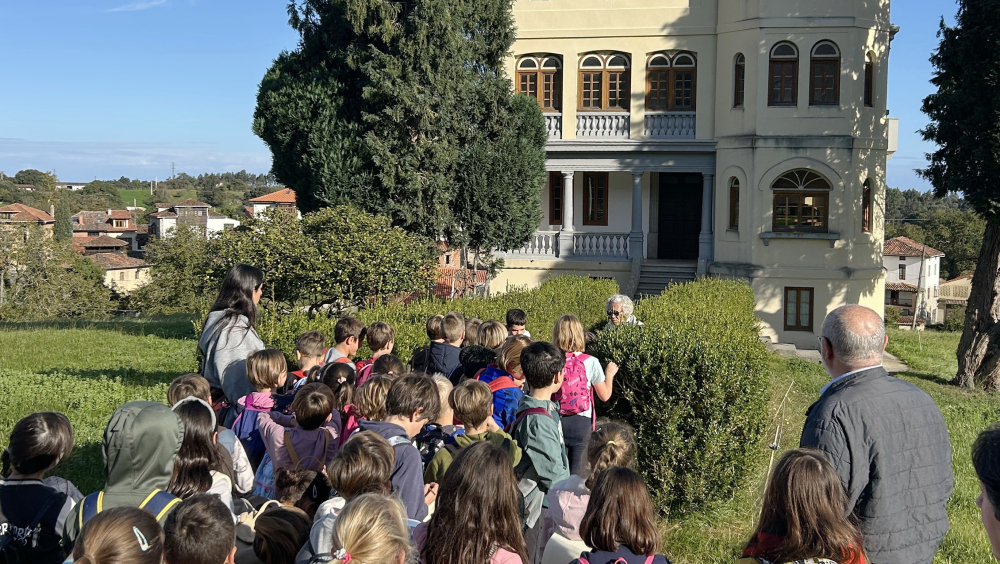 Un grupo de niñas y niños con mochilas entrando en el edificio El Castillo (casona) para una visita guiada. Están en una zona verde al aire libre, rodeada de árboles. El edificio tie-ne una torre redonda, ventanas arqueadas y tejado de tejas.