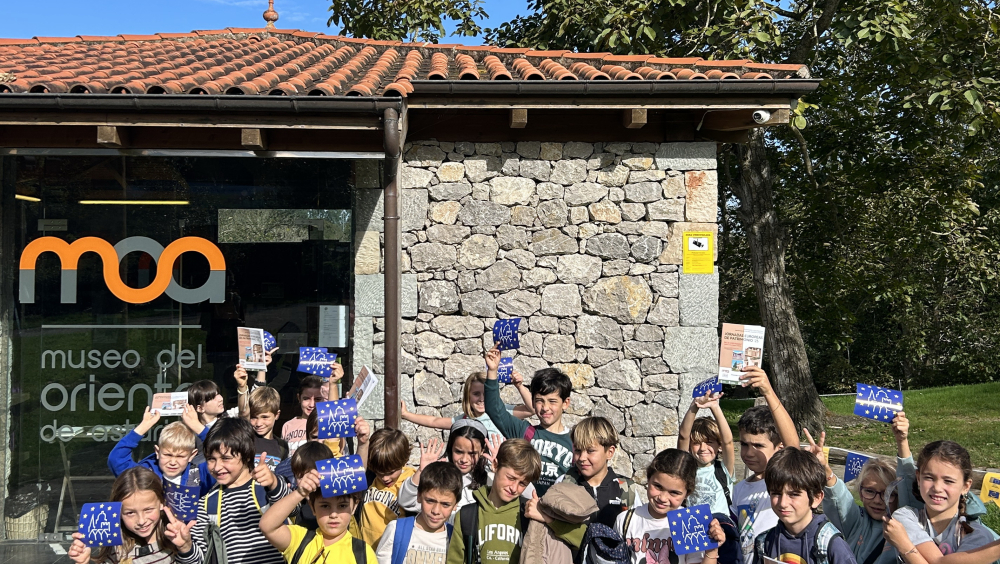 Un grupo de niños y niñas sonrientes está reunido a la entrada del Museo del Oriente de Asturias. Sostienen cuadernillos con las actividades didácticas de «Casonas y Casinas», así como pega-tinas de las JEP. El entorno es verde y natural.