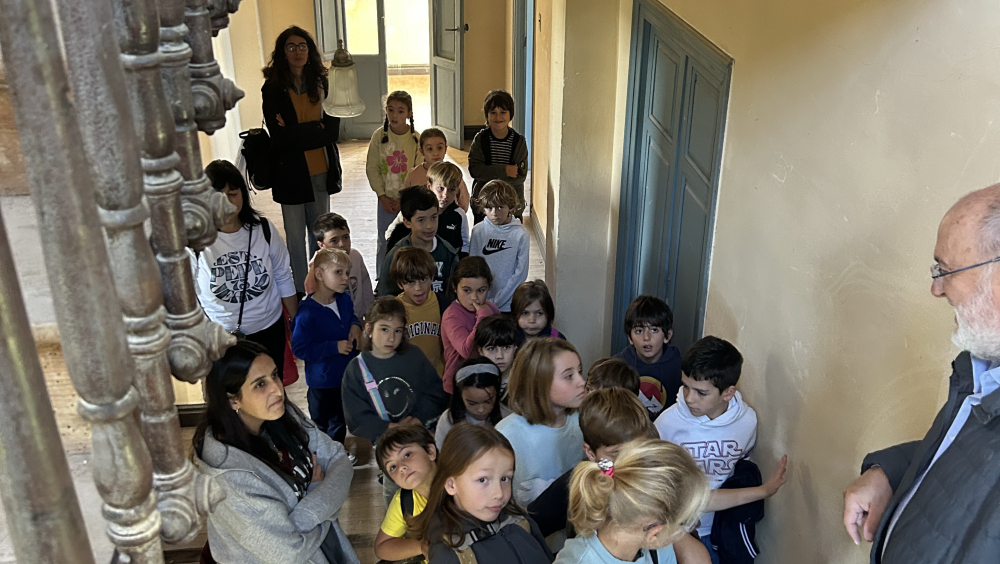 En una amplia escalera interior de madera, un grupo de niñas y niños escucha con atención a un hombre mayor que les habla. Están participando en la visita guiada al edificio histórico de El Castillo (casona). El ambiente es cálido y lleno de detalles arquitectónicos.