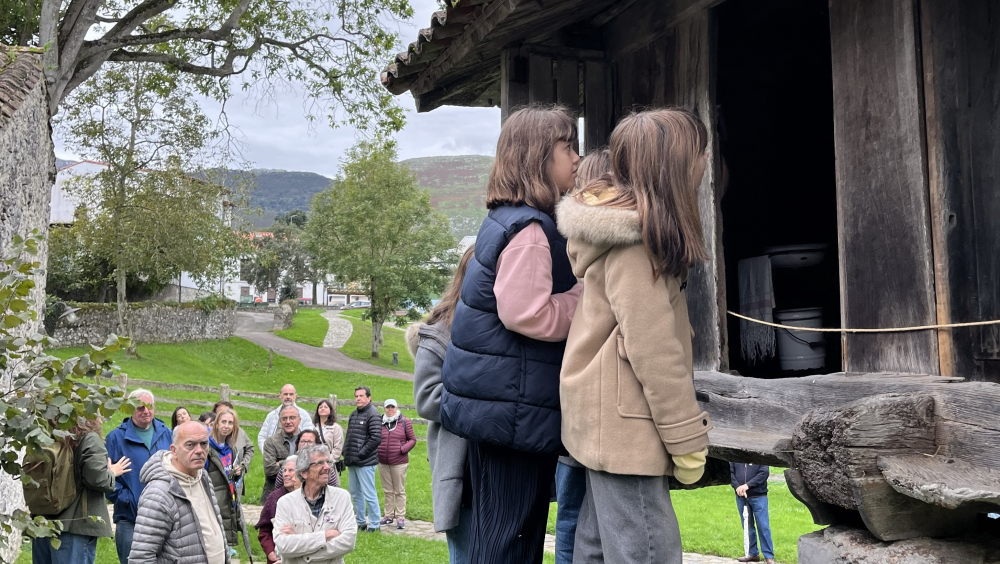 Varias personas, incluidos niños y adultos, rodean un hórreo (construcción de madera elevada sobre pilares de piedra). Dos niñas subidas en una escalera de pieza maciza miran curiosas hacia el interior del hórreo. El grupo participa en una visita cultural guiada al aire libre, en un entorno rural con hierba, árboles y colinas al fondo.