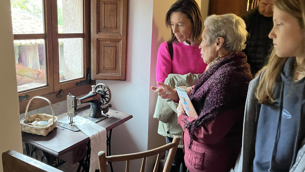 Varias personas observan una antigua máquina de coser Singer colocada sobre una mesa junto a una ventana. Una mujer mayor señala la máquina explicando su uso o historia a los miembros más jóvenes del grupo. Junto a la máquina hay una cesta con hilos y telas. Se encuentran en el interior de una casina, en un ambiente cálido y rústico.