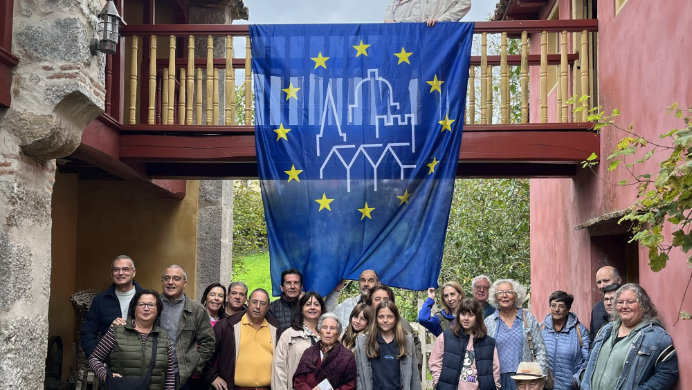 Un grupo de personas está reunido frente a un edificio antiguo con un pasadizo volado de madera y paredes de piedra. Del pasadizo cuelga una gran bandera con el logotipo de las JEP. Dos personas observan desde arriba, mientras el grupo permanece en un patio empedrado, como parte de la visita guiada a la Casa de Llacín.