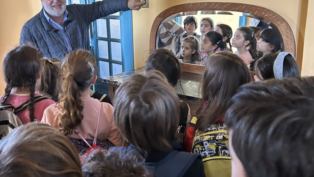 Dentro de un comedor con paredes amarillas y puerta acristalada azul, un hombre mayor explica la historia del edificio a un grupo de escolares que le escuchan con atención. Algunos llevan mochilas. Un gran espejo refleja sus caras, capturando el momento de aprendizaje y curiosidad compartida.