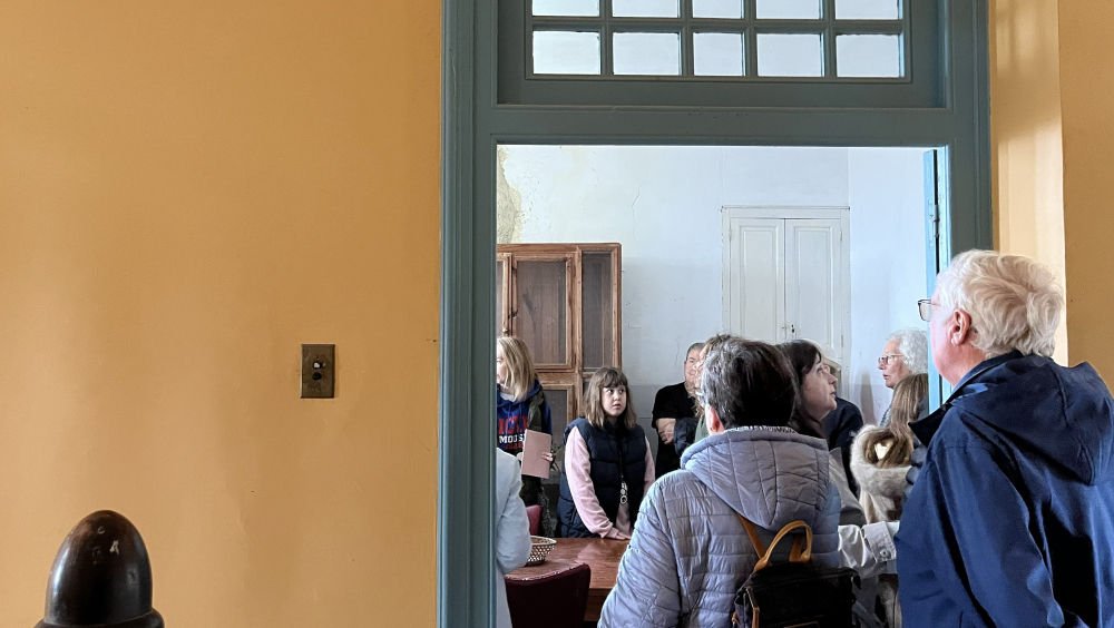 Desde un distribuidor con pared amarilla, se ve a través de una puerta azul acristalada a varias personas reunidas en una amplia cocina. Están alrededor de una mesa, atendiendo a la explicación de una visita guiada al interior del edificio histórico El Castillo.
