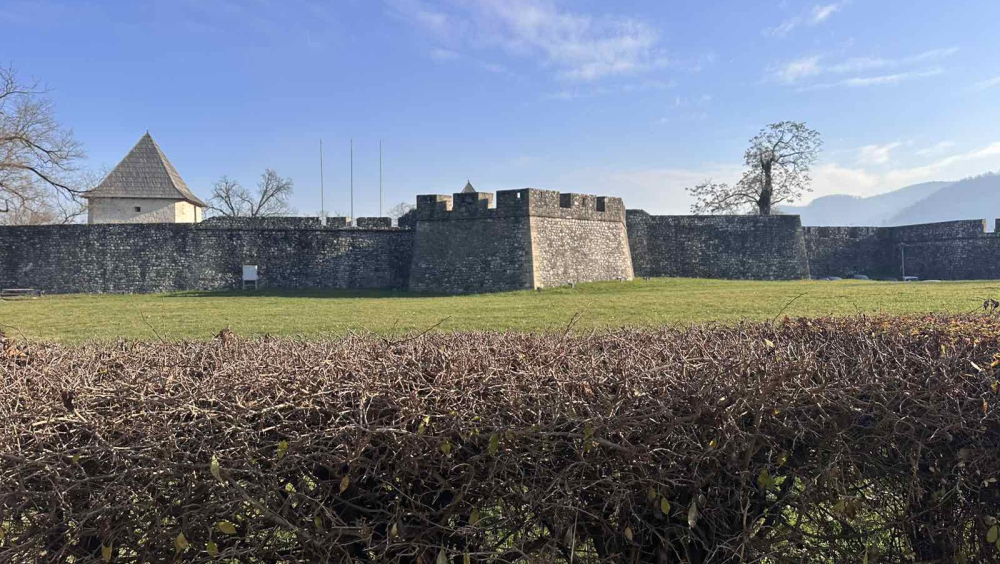 Wide view of Kastel Fortress in Banja Luka, showing thick stone walls and towers surrounded by a green grassy area under a clear blue sky.