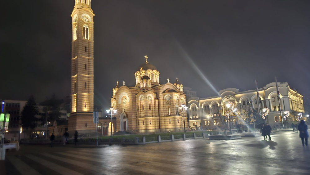 Night view of the Cathedral of Christ the Saviour in Banja Luka, an illuminated Orthodox church with a tall bell tower, located on a city square with a pedestrian area and surrounding buildings.