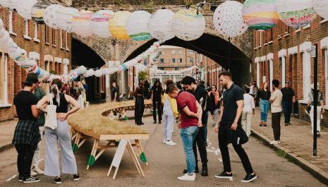 A view looking down Beck Road, towards the railway arch, the Victorian terrace houses running parallel to each other either side of the frame. Between them, Beyond Beck Road visitors are looking at the architecture, at hanging paper lanterns overhead, and at Luke Deering's art installation 'Reclaim the Commons': a meadow elevated on a tabletop running off into the distance. 