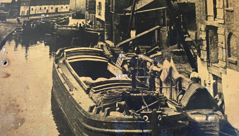 An old photo of Preston Brook wharf. In the foreground 3 men are working a crane unloading sacks from a barge into a narrowboat tied alongside. Behind them two barges are being maneuvered into a warehouse. 