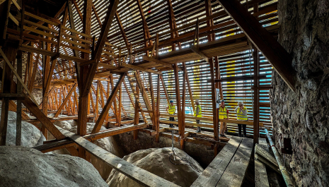Workers in bright yellow safety vests inspect a complex wooden roof framework under renovation inside large open room. They are walking on wooden walkways built over large rocks forming the floor.