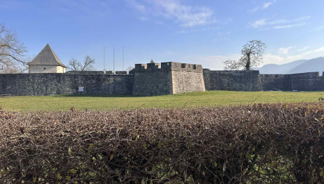 Wide view of Kastel Fortress in Banja Luka, showing thick stone walls and towers surrounded by a green grassy area under a clear blue sky.