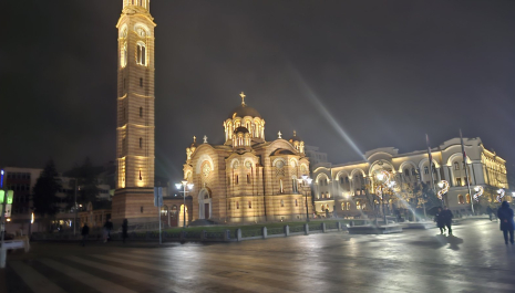 Night view of the Cathedral of Christ the Saviour in Banja Luka, an illuminated Orthodox church with a tall bell tower, located on a city square with a pedestrian area and surrounding buildings.