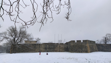Winter view of Kastel Fortress in Banja Luka, with snow covering the ground, bare tree branches overhead, and two children standing in front of the stone walls.