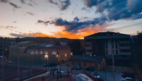 Pogled na Sokolski dom i sportsko igralište u Banjoj Luci u sumrak, sa djecom koja igraju košarku, okružen zgradama grada i obojenim nebom u zalasku sunca.